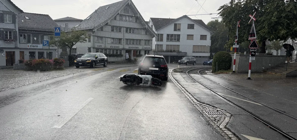 Ein Motorrad ist auf einer regennassen Straße umgestürzt und liegt vor einem Auto. Der Unfall ereignete sich in einem Stadtgebiet mit altertümlichen Gebäuden.