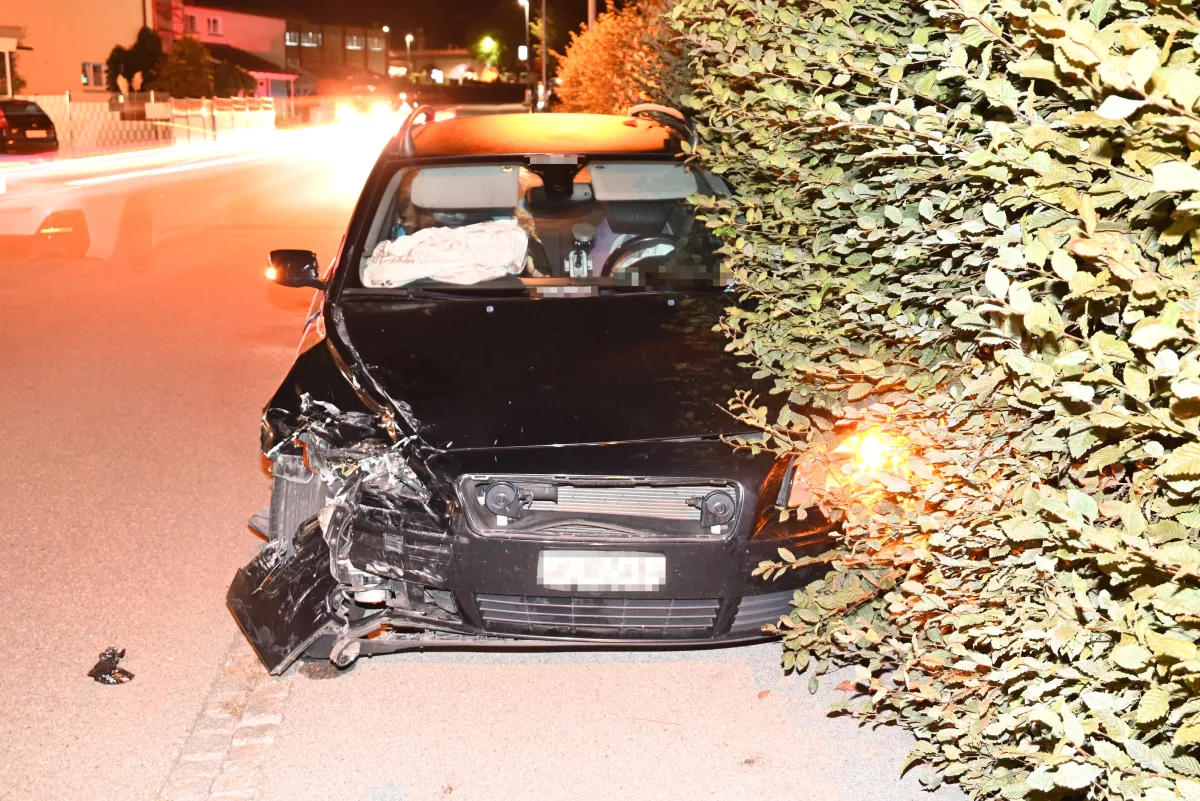 Ein schwarzer PKW mit beschädigter Front steht nachts an einer Hecke. Das Fahrzeug ist durch einen Unfall in Mitleidenschaft gezogen.