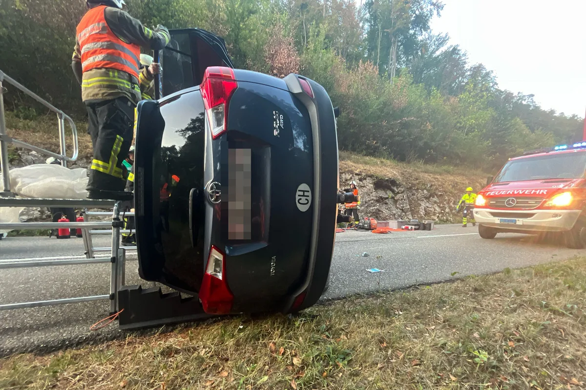 Ein Auto liegt auf der Seite einer Landstraße, während Feuerwehrleute es sichern. Daneben ein Feuerwehrfahrzeug mit Blaulicht.