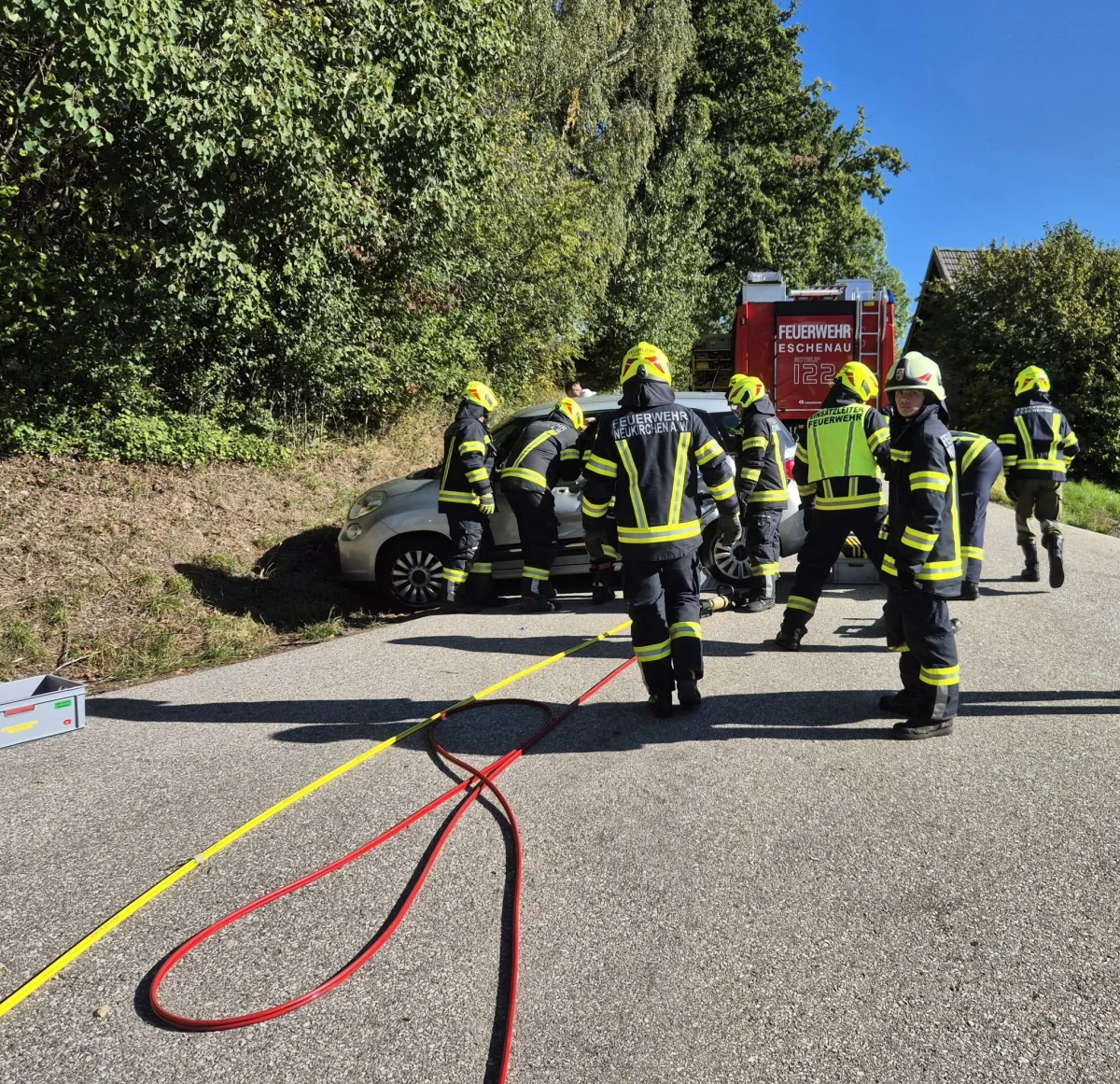 Mehrere Feuerwehrleute arbeiten an einem silbernen Auto, das am Straßenrand steht. Ein Feuerwehrfahrzeug ist im Hintergrund sichtbar.