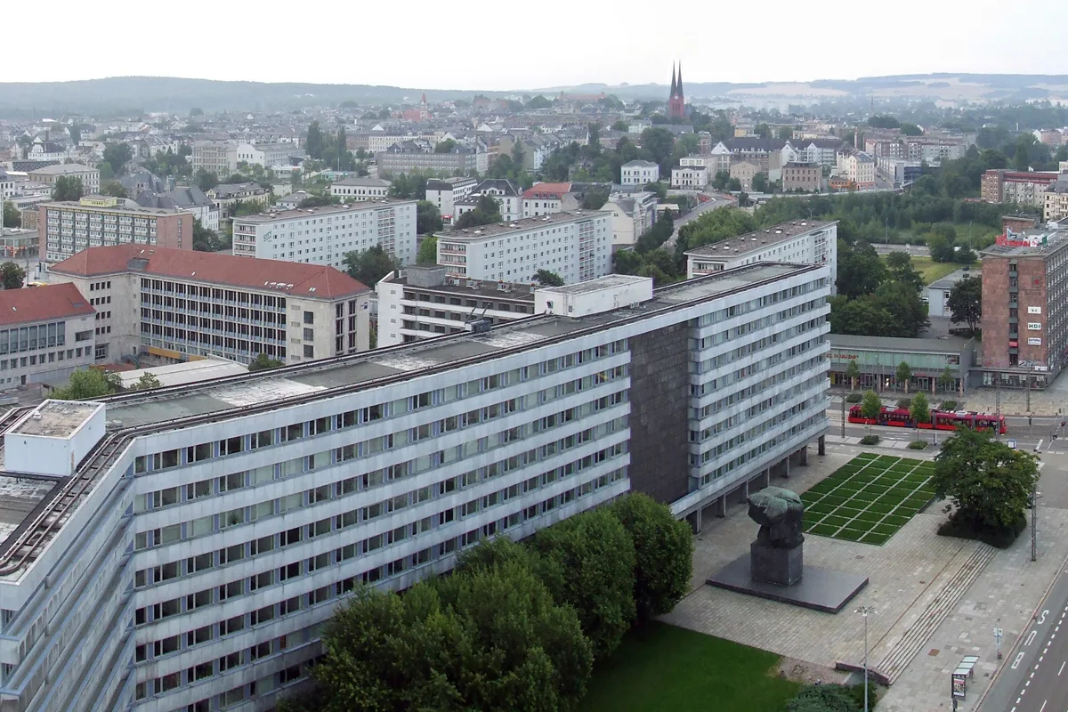 Eine aus der Vogelperspektive aufgenommene Stadtansicht zeigt einen großen Wohnblock im Vordergrund und eine weitläufige urbane Umgebung im Hintergrund.