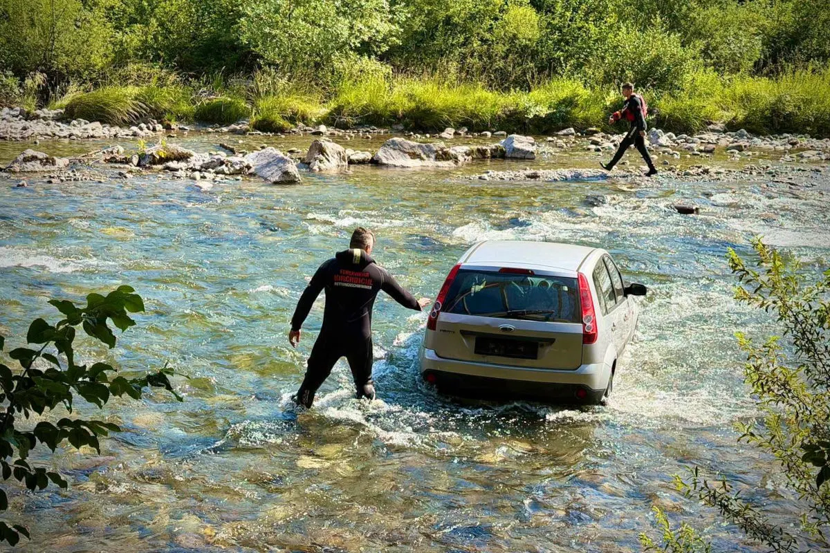 Ein silbernes Auto befindet sich im flachen Wasser eines Flusses. Einsatzkräfte der Wasserrettung stehen ebenfalls im Fluss und bewegen sich um das Fahrzeug herum.
