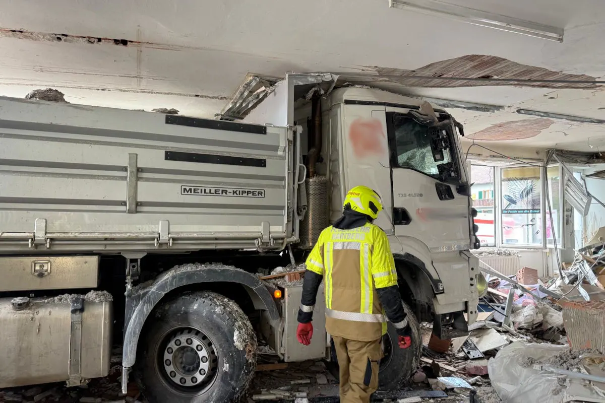 Ein Lkw ist in den Innenraum eines Gebäudes gefahren. Ein Feuerwehrmann in gelber Schutzkleidung prüft die Situation im zerstörten Bereich.