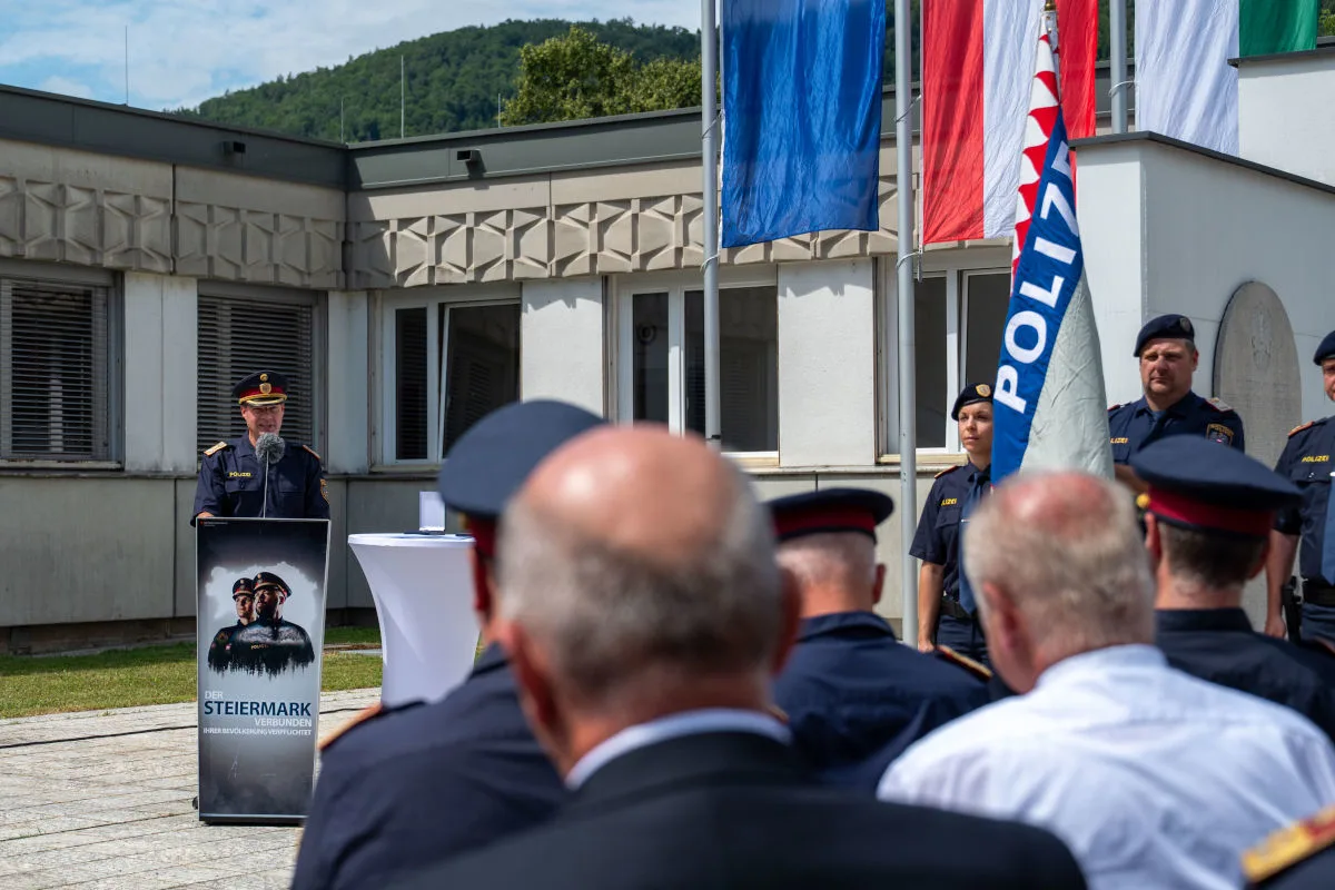 Ein Polizist steht an einem Rednerpult mit Steiermark-Logo und spricht zu einer Gruppe von Kollegen. Einige Polizeiflaggen sind sichtbar.
