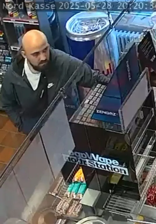 Ein glatzköpfiger Mann mit Bart ist an der Kasse eines Geschäfts zu sehen, von einer Überwachungskamera aufgenommen.