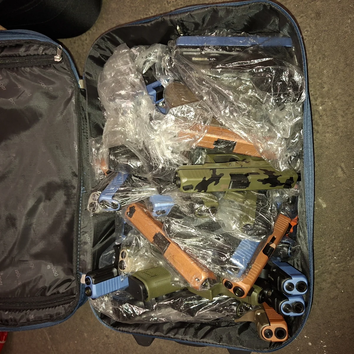 Ein offener Koffer enthält zahlreiche Schusswaffen, die in Plastikfolie verpackt sind. Der Koffer liegt auf einem Bodenbelag und die Pistolen sind in verschiedenen Farben und Mustern dargestellt.
