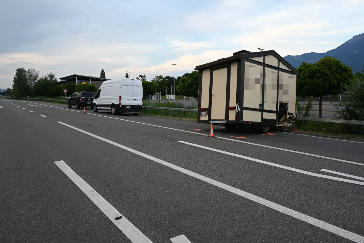 Ein beige-brauner Anhänger mit sichtbaren Schäden steht am Straßenrand, daneben ein weißer Van und ein weiteres Fahrzeug.