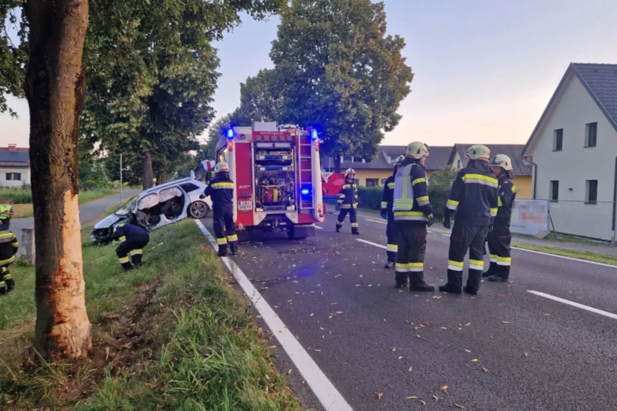 Ein Auto steht stark beschädigt neben einem Baum mit sichtbaren Rindenverletzungen. Feuerwehrleute sind vor Ort und sichern die Unfallstelle ab.