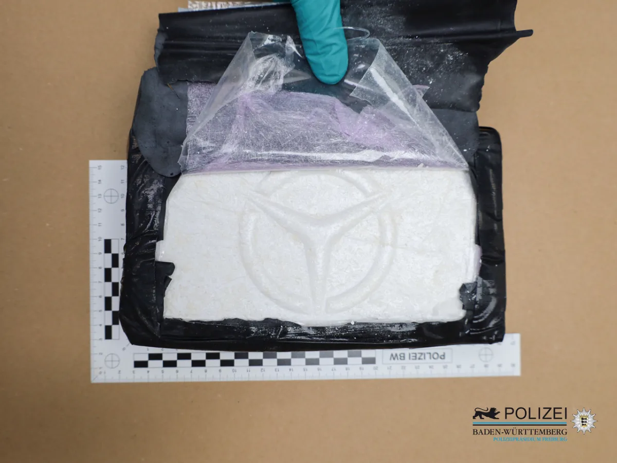 Ein rechteckiges, weißes Paket mit einem eingeprägten Symbol wird teilweise von schwarzem Kunststoff umgeben. Eine Person in Handschuhen entfernt Plastik.