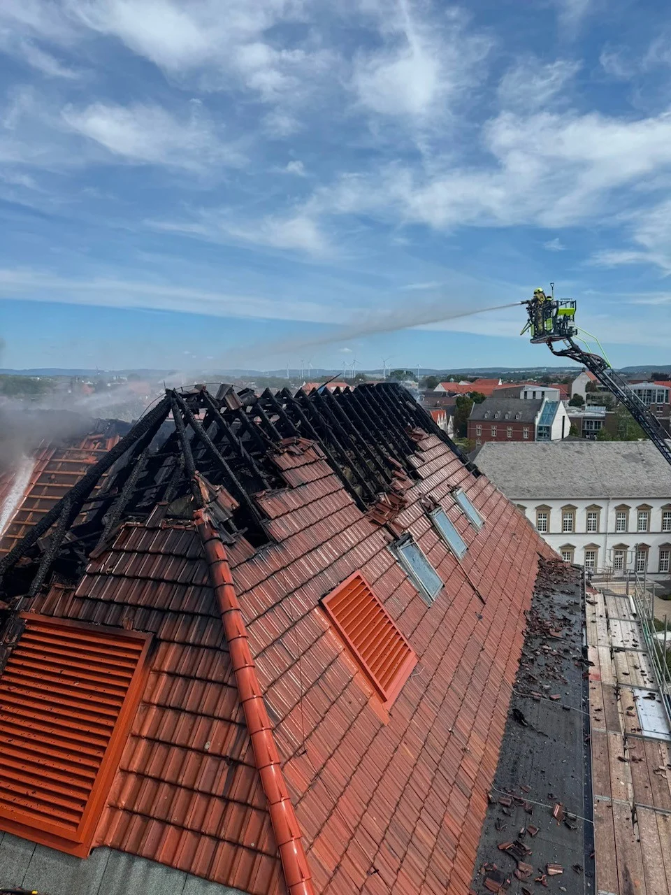 Ein teilweise zerstörtes Gebäudedach wird von Feuerwehrleuten von einer Leiter aus gelöscht. Der Himmel ist klar.