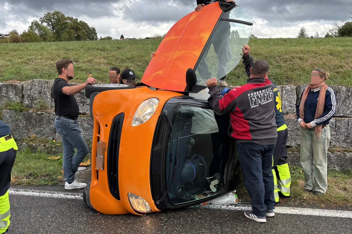 Ein orangefarbener Kleinwagen liegt auf der Seite neben einer Straße. Menschen stehen um das Fahrzeug herum.