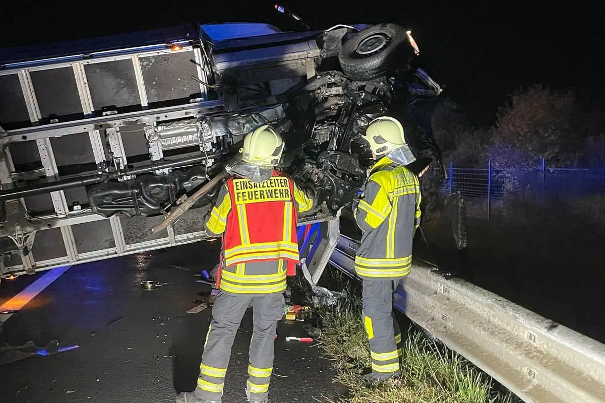 Zwei Feuerwehrleute in Schutzkleidung stehen vor einem seitlich umgekippten LKW auf einer Straße in der Nacht.