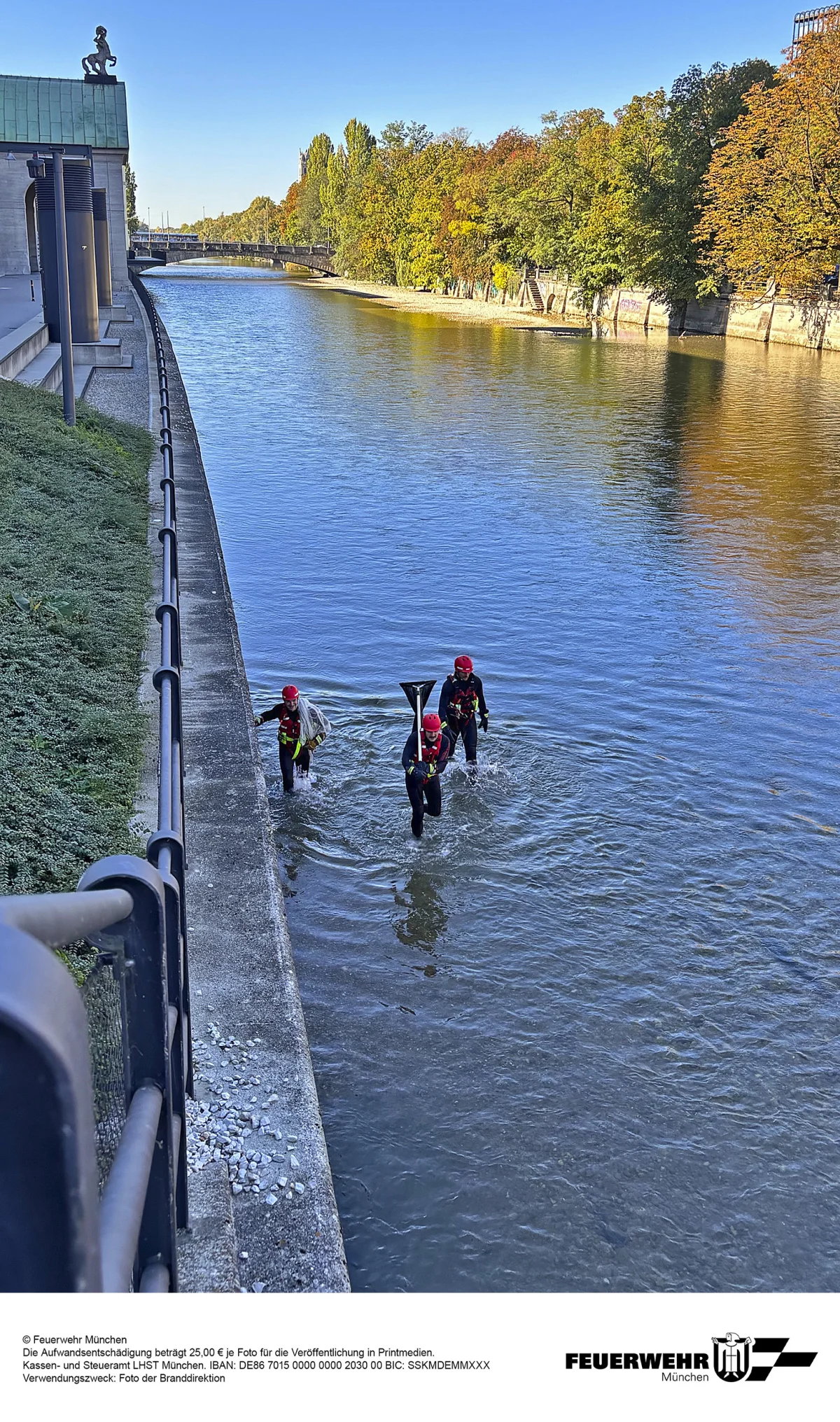 Drei Feuerwehrleute mit Helmen und Einsatzkleidung waten durch einen Fluss, nahe einer betonierten Ufermauer.