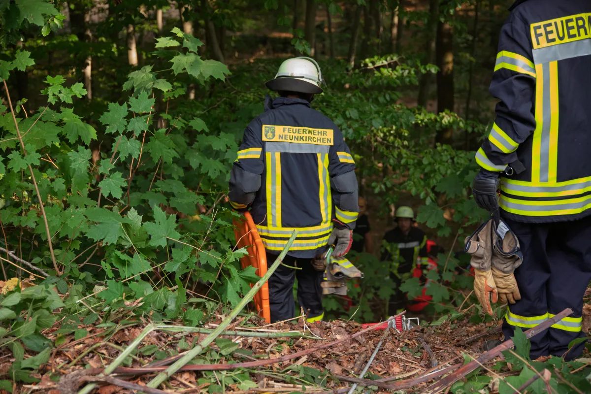 Mehrere Feuerwehrleute in Schutzkleidung stehen in einem bewaldeten Gebiet. Einer trägt Ausrüstung, während andere im Hintergrund zu sehen sind.