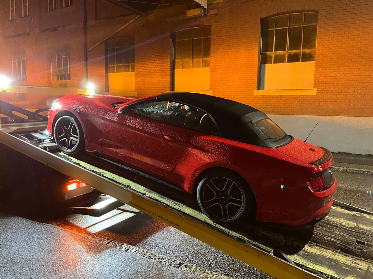 Ein rotes Cabriolet steht bei Nacht im Regen auf einem Abschleppwagen vor einem Backsteingebäude. Die Straße ist nass.