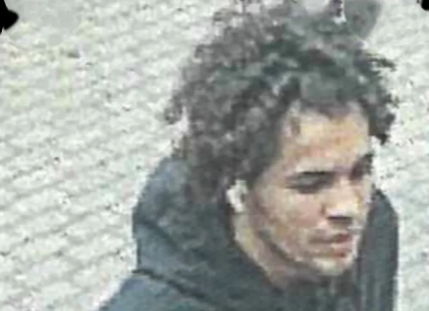 Ein Mann mit lockigem Haar und Ohrhörern ist auf einem gepflasterten Untergrund zu sehen. Er trägt einen dunklen Hoodie.