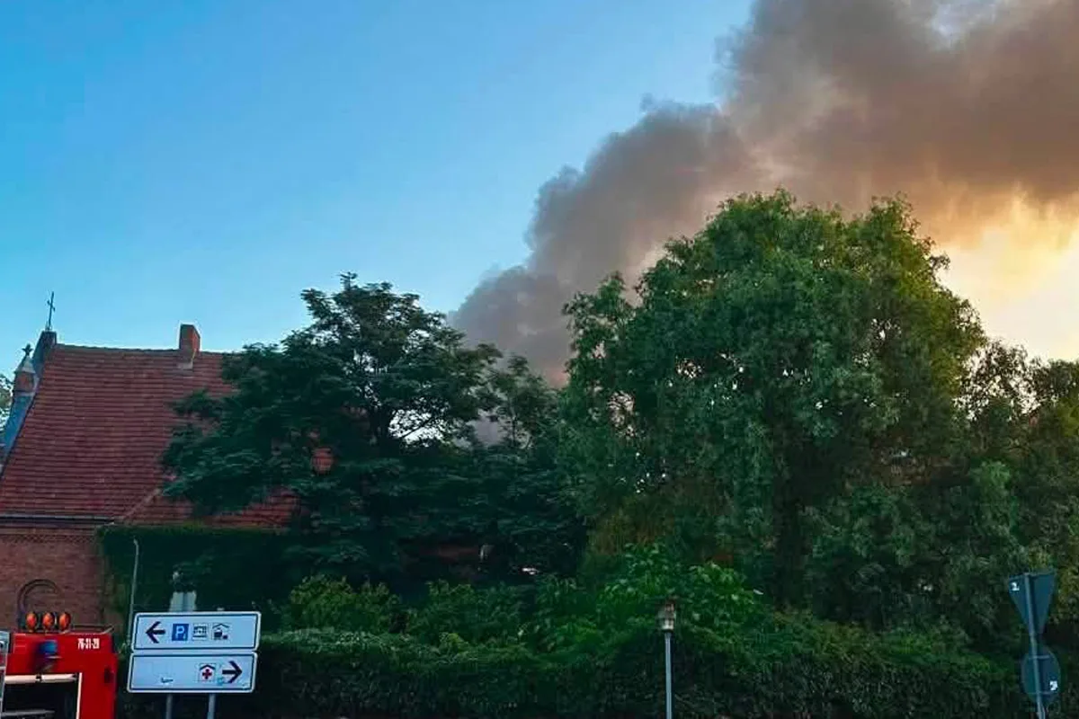 Eine große dunkle Rauchwolke erhebt sich über einem Haus und Bäumen. Im Vordergrund ist ein Teil eines Feuerwehrfahrzeugs sichtbar.
