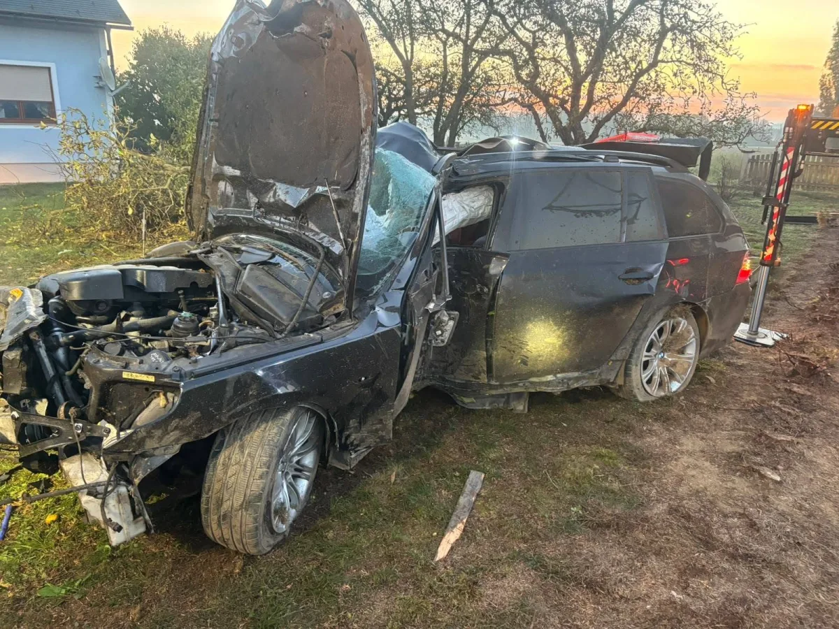 Ein schwarzes Auto mit schwerem Frontschaden steht mit geöffneter Motorhaube auf einer ländlichen Wiese. Der Sonnenuntergang ist im Hintergrund zu sehen.