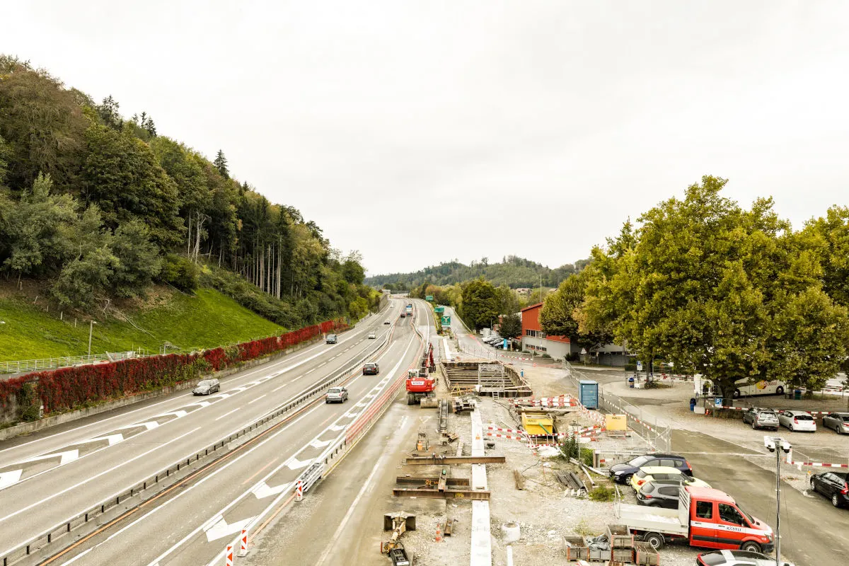 Eine Baustelle befindet sich neben einer Autobahn, mit mehreren Fahrspuren und vorbeifahrenden Autos. Am Straßenrand stehen Bäume und es sind Baustellenfahrzeuge sichtbar.