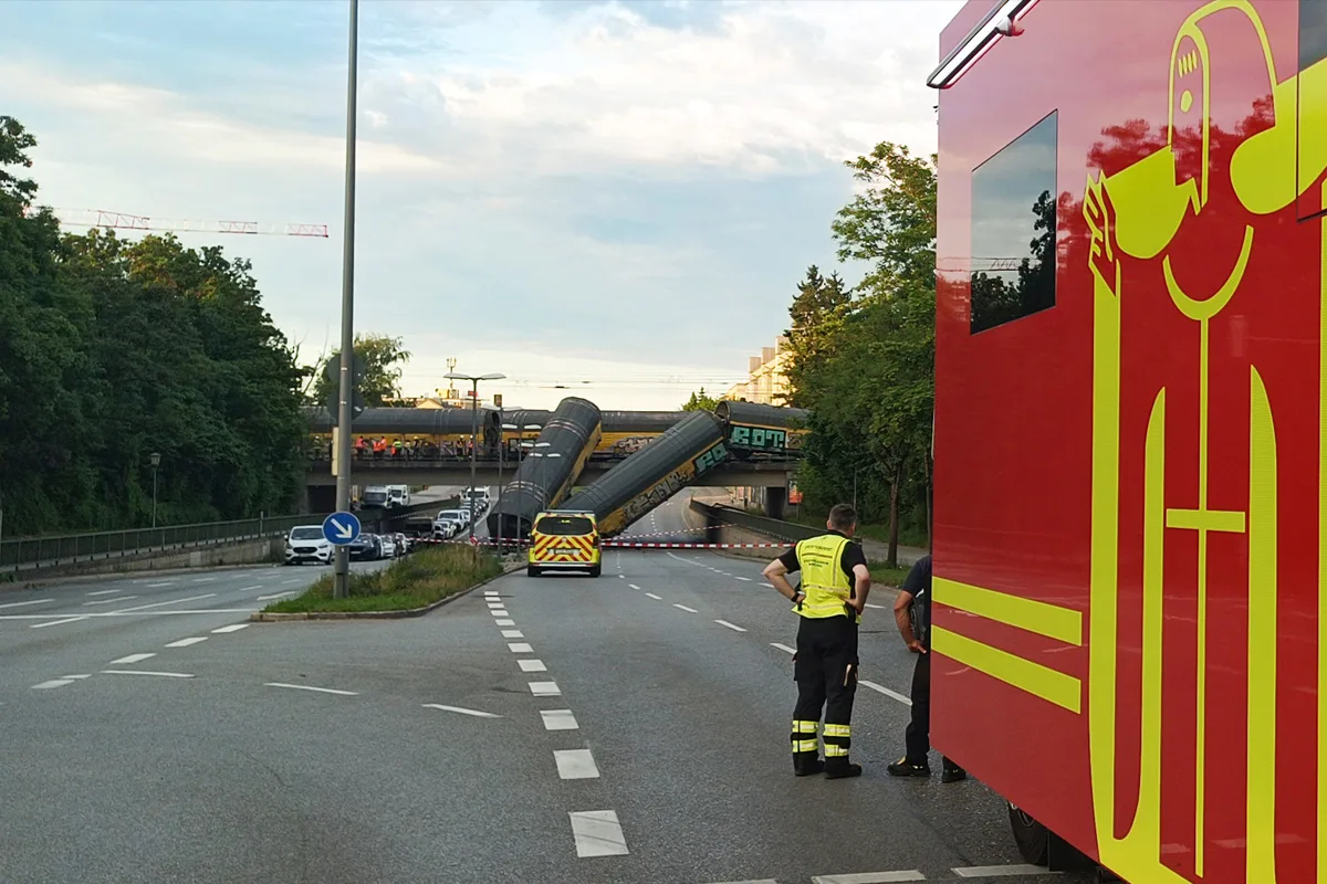 Ein umgestürzter Lkw-Anhänger liegt auf einer Brücke und blockiert den Verkehrsfluss auf der darunterliegenden Straße. Einsatzfahrzeuge sind vor Ort.