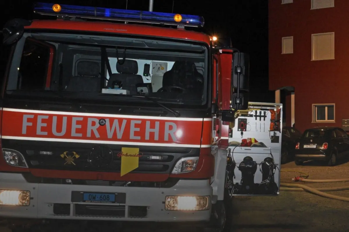Ein rotes Feuerwehrfahrzeug mit der Aufschrift 'FEUERWEHR' ist zu sehen. Der hintere Bereich des Fahrzeugs ist offen, und ein Wohngebäude ist im Hintergrund sichtbar.