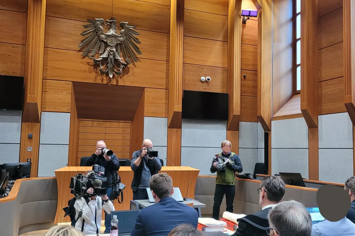 Mehrere Fotografen im Gerichtssaal richten ihre Kameras auf eine Personengruppe. Der Raum ist holzvertäfelt und mit dem Bundesadler dekoriert.