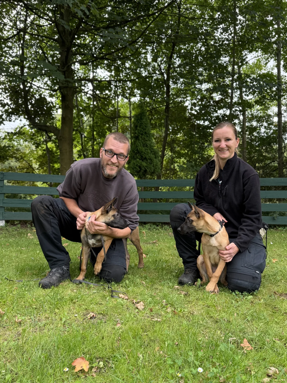Zwei Personen in dunkler Kleidung knien auf einer Wiese mit jeweils einem jungen Hund. Im Hintergrund sind Bäume zu sehen.