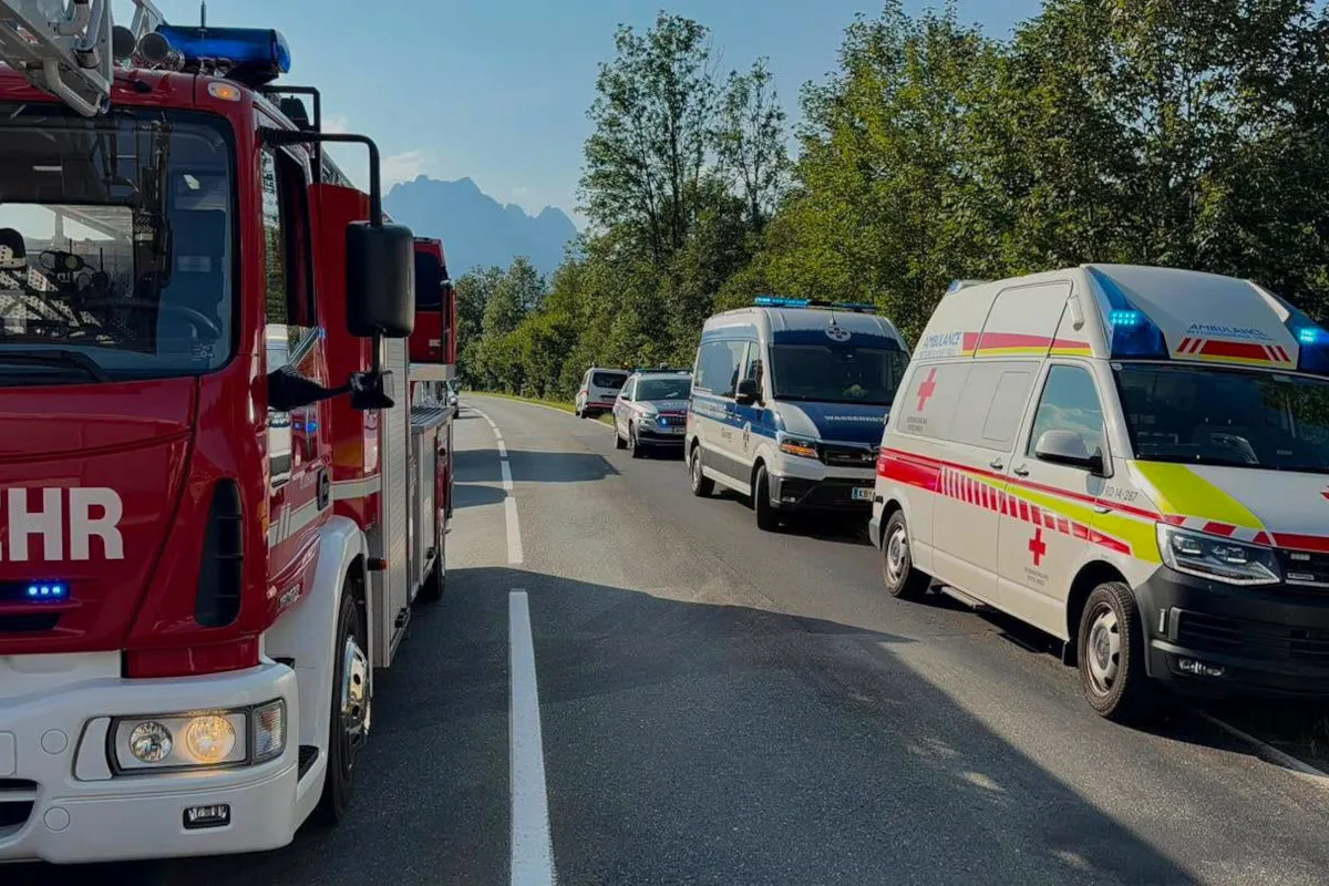 Mehrere Rettungsfahrzeuge, einschließlich eines Feuerwehrautos und eines Krankenwagens, stehen auf einer einsamen Straße in einer grünen, ländlichen Gegend; Berge im Hintergrund.