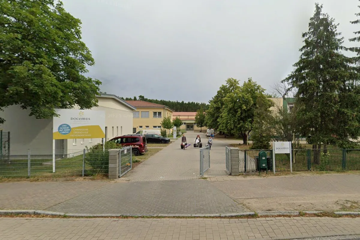 Der Eingang eines Gymnasiums mit parkendem Auto und drei Personen, die eine breite gepflasterte Einfahrt entlang gehen. Links und rechts der Einfahrt befinden sich Bäume und Schulgebäude.