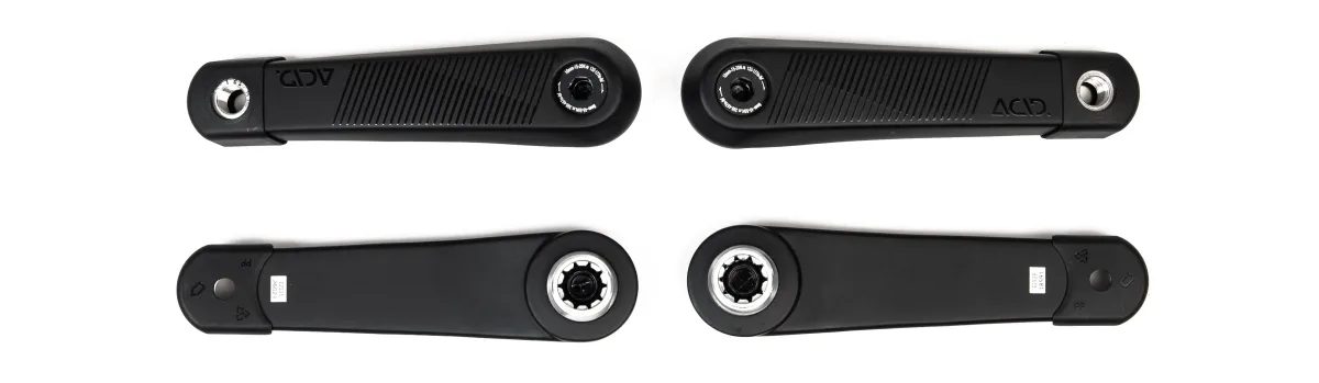 Das Bild zeigt zwei schwarze Fahrradkurbeln, die nebeneinander horizontal liegen. Beide Kurbeln haben metallische Einsätze an den Enden.