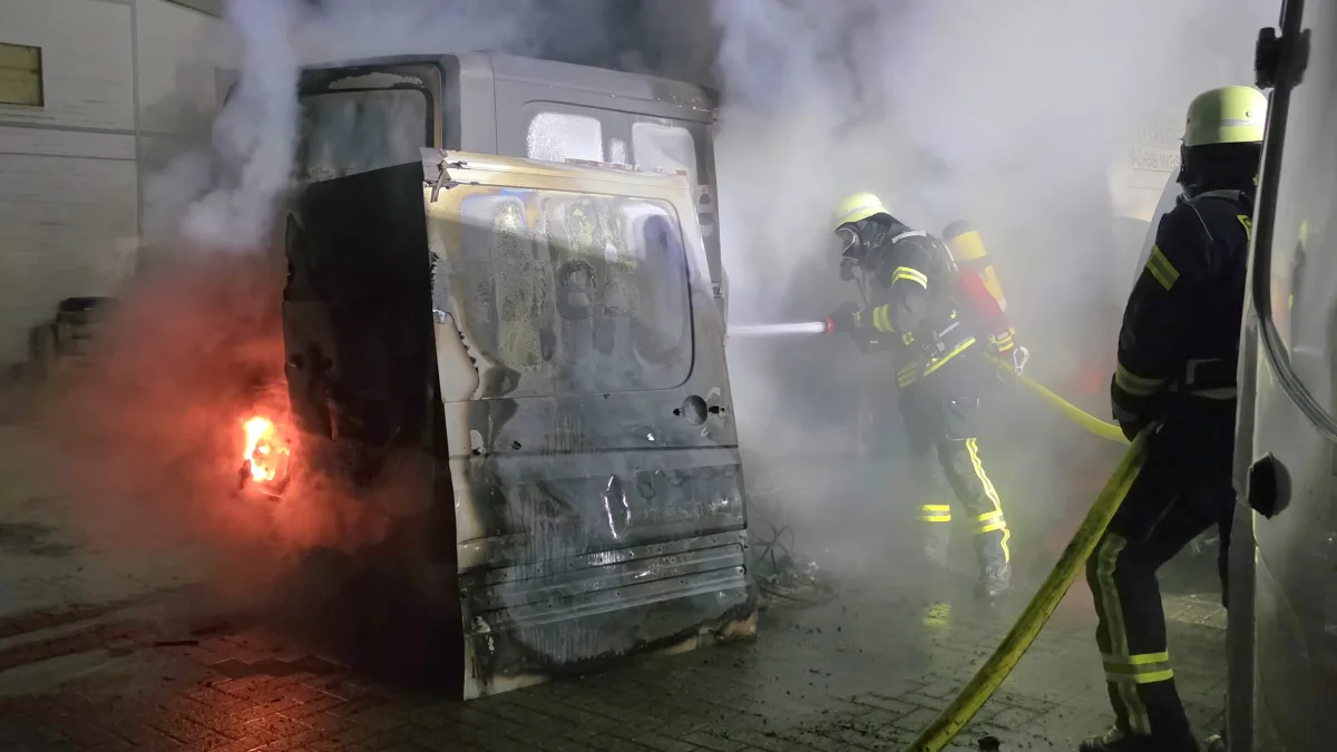 Zwei Feuerwehrmänner bekämpfen ein Feuer in einem stark beschädigten Fahrzeug. Dichte Rauchschwaden umhüllen die Szene.