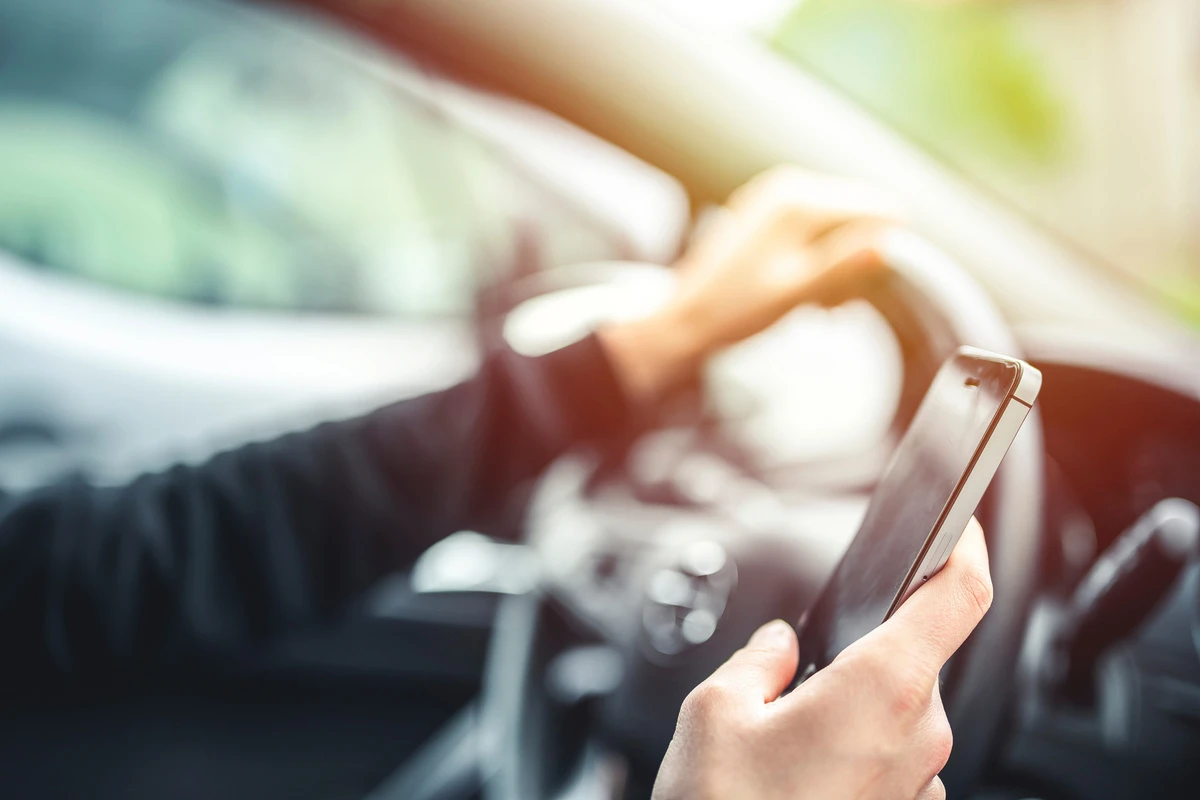 Eine Person hält ein Smartphone in der rechten Hand, während sie mit der linken das Lenkrad eines Autos steuert. Sonnenlicht fällt auf die Szene.