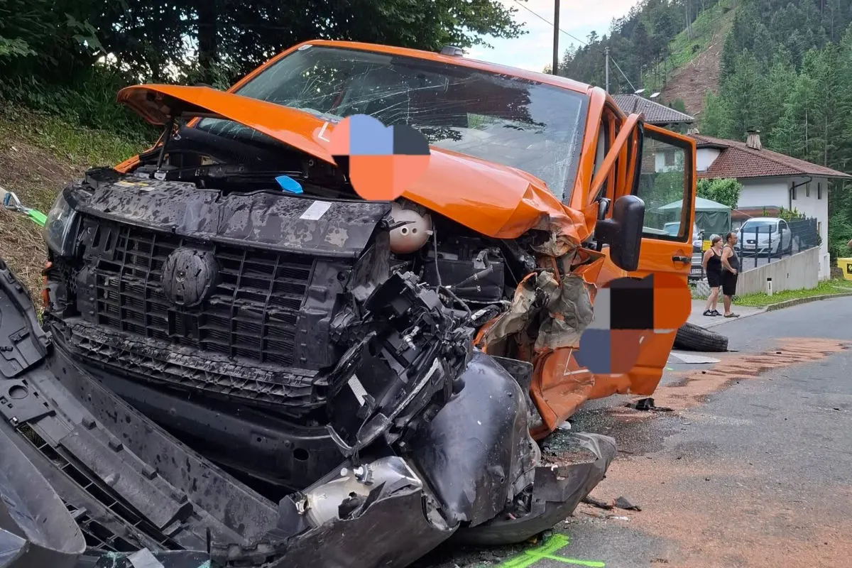 Ein orangefarbener Transporter mit schwer demolierter Front steht am Straßenrand. Im Hintergrund sind zwei Personen zu sehen.