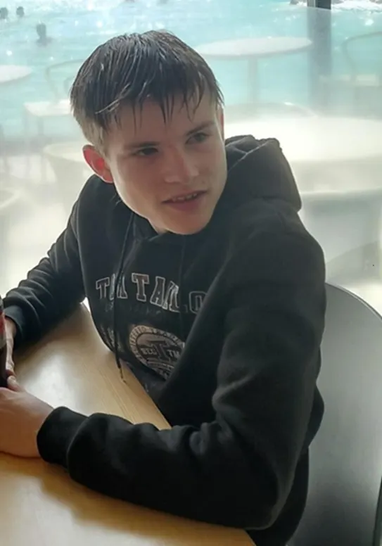 Ein junger Mann mit nassem Haar trägt einen dunklen Hoodie. Er sitzt an einem Tisch, der Hintergrund ist verschwommen und lichtdurchflutet.