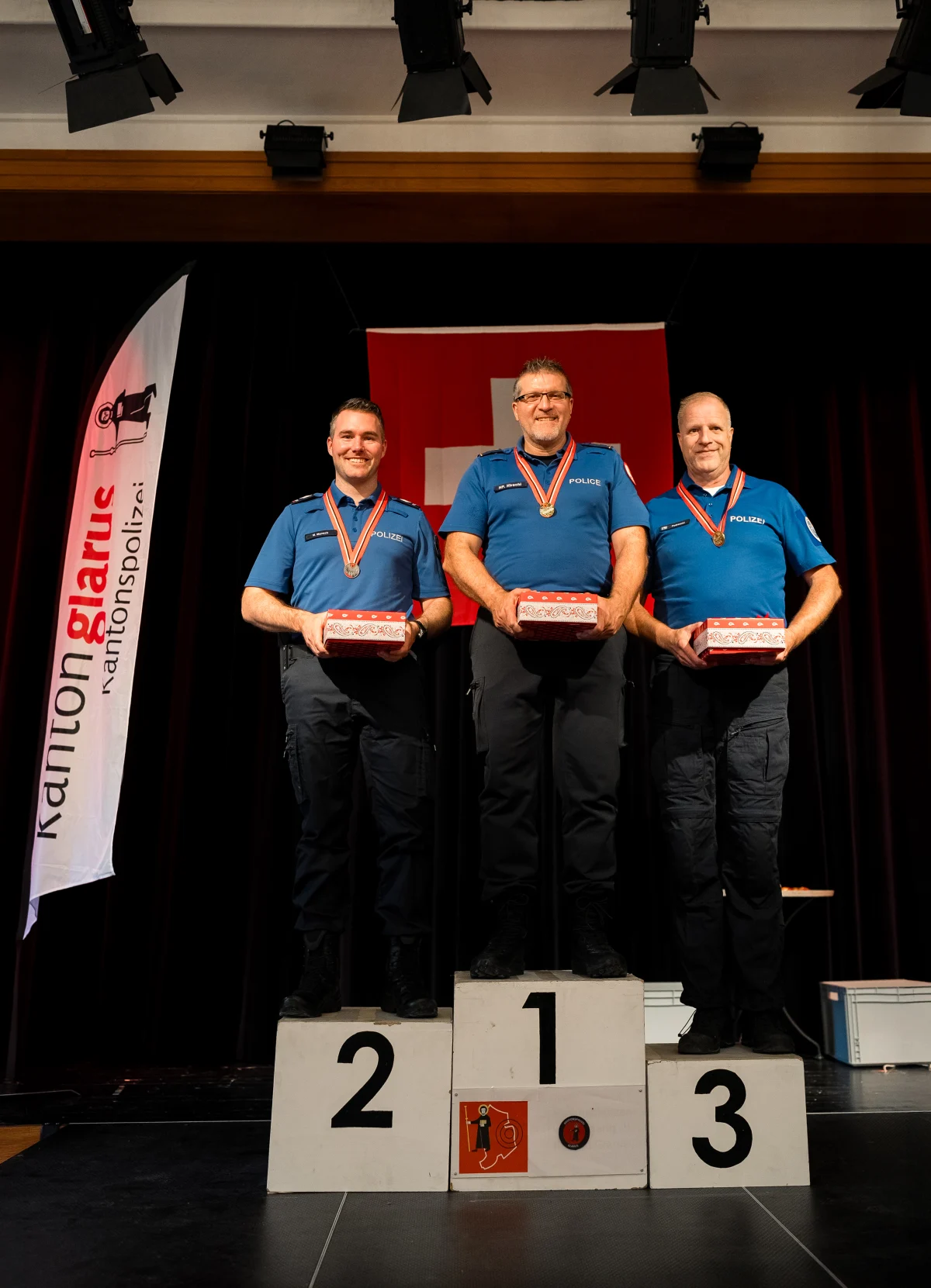 Drei Männer in blauer Polizeiuniform stehen auf einem Siegerpodest mit Medaillen um den Hals und halten Geschenke.