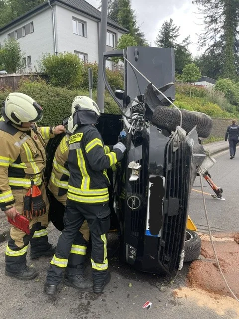 Zwei Feuerwehrleute in Uniform arbeiten an einem umgestürzten Fahrzeug, das auf der Seite liegt, in einer Wohnstraße.