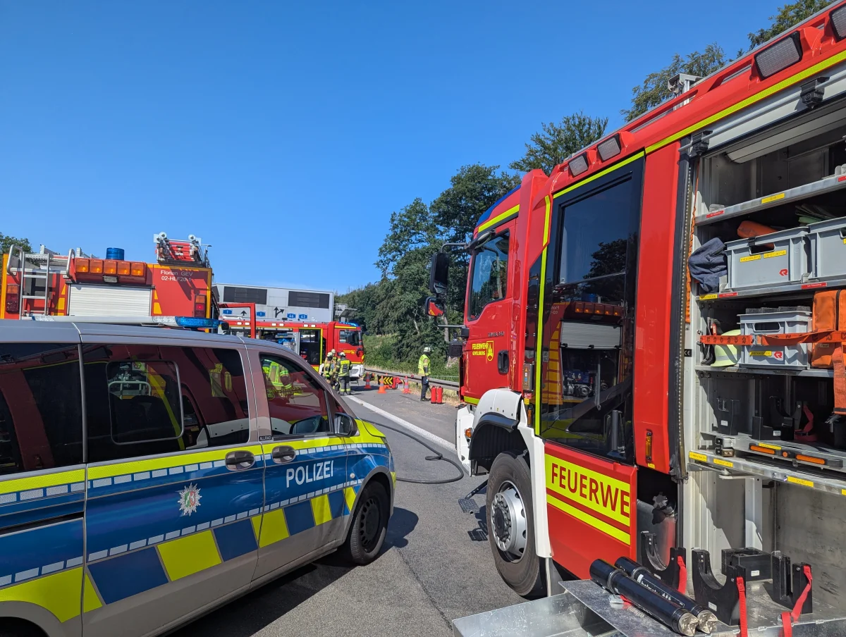 Ein Polizeifahrzeug und mehrere Feuerwehrfahrzeuge stehen nebeneinander auf einer Straße. Im Hintergrund sind Bäume zu sehen.