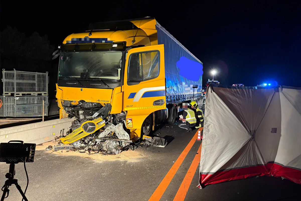 Ein gelber Lastwagen mit stark beschädigter Front auf einer Autobahn bei Nacht. Zwei Einsatzkräfte arbeiten nahe dem Fahrzeug.