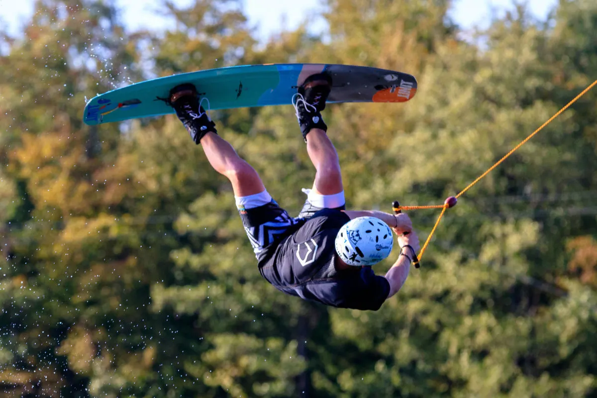 Ein Wakeboarder mit Helm und Schwimmweste springt hoch in der Luft. Im Hintergrund sind unscharfe Bäume zu sehen und die Sonne scheint.