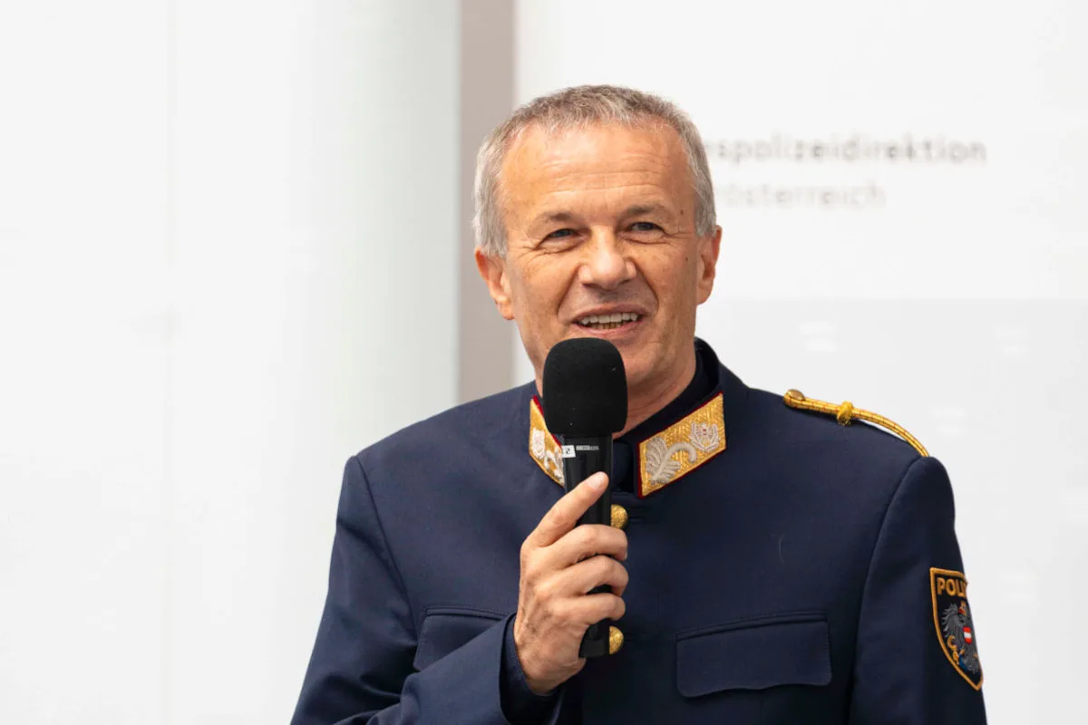 Ein Mann in einer dunkelblauen Polizeiuniform spricht in ein Mikrofon. Das Abzeichen auf dem Ärmel hat den Schriftzug 'Polizei'.