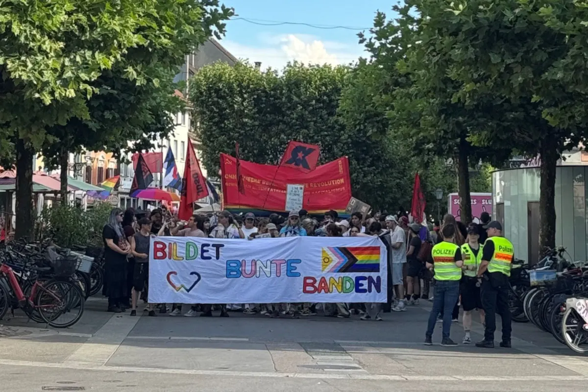 Eine Gruppe von Menschen hält bei einer Demonstration ein buntes Transparent mit Aufschrift vor sich. Polizeibeamte beobachten die Szene seitlich.