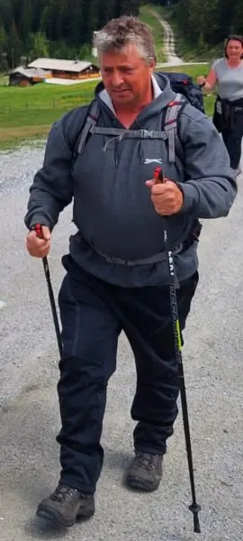 Ein Mann in Outdoor-Kleidung geht mit Trekkingstöcken einen Schotterweg entlang, im Hintergrund eine ländliche Landschaft.