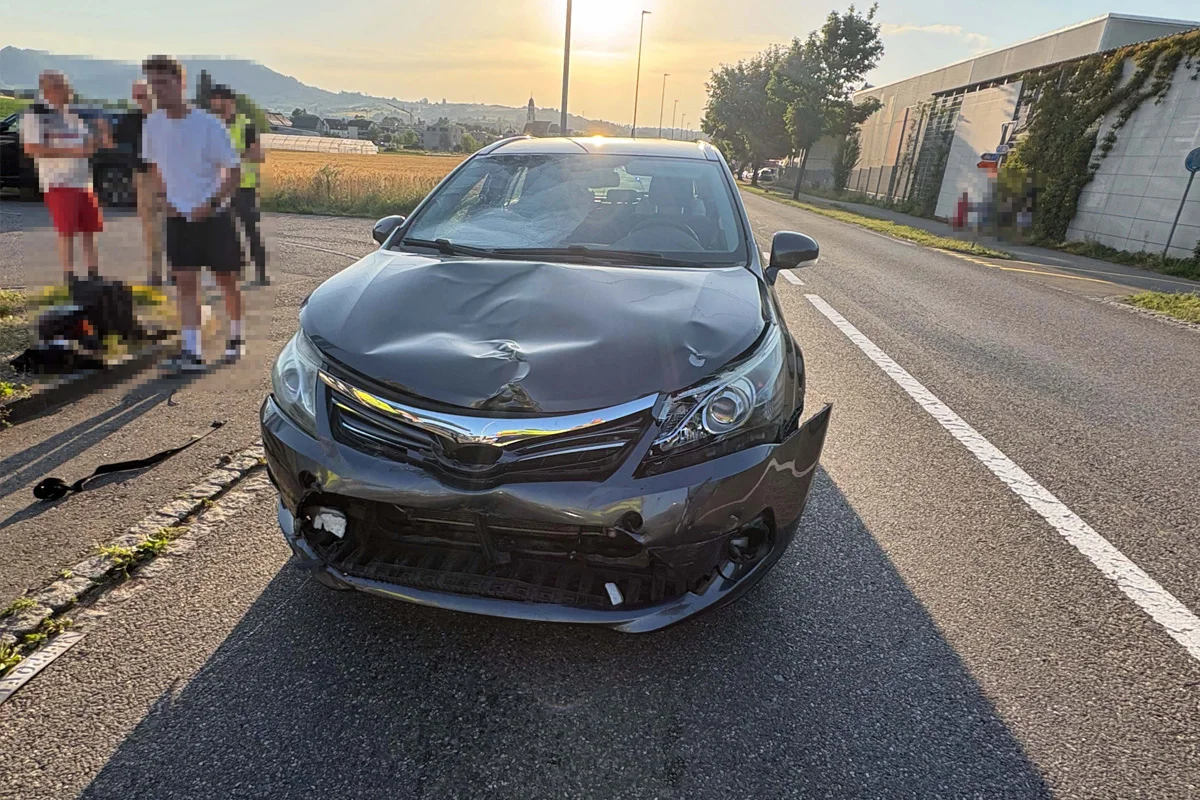 Ein Auto mit stark beschädigter Front steht auf einer Straße. Im Hintergrund sind Felder und Gebäude zu sehen, Menschen befinden sich am Straßenrand.