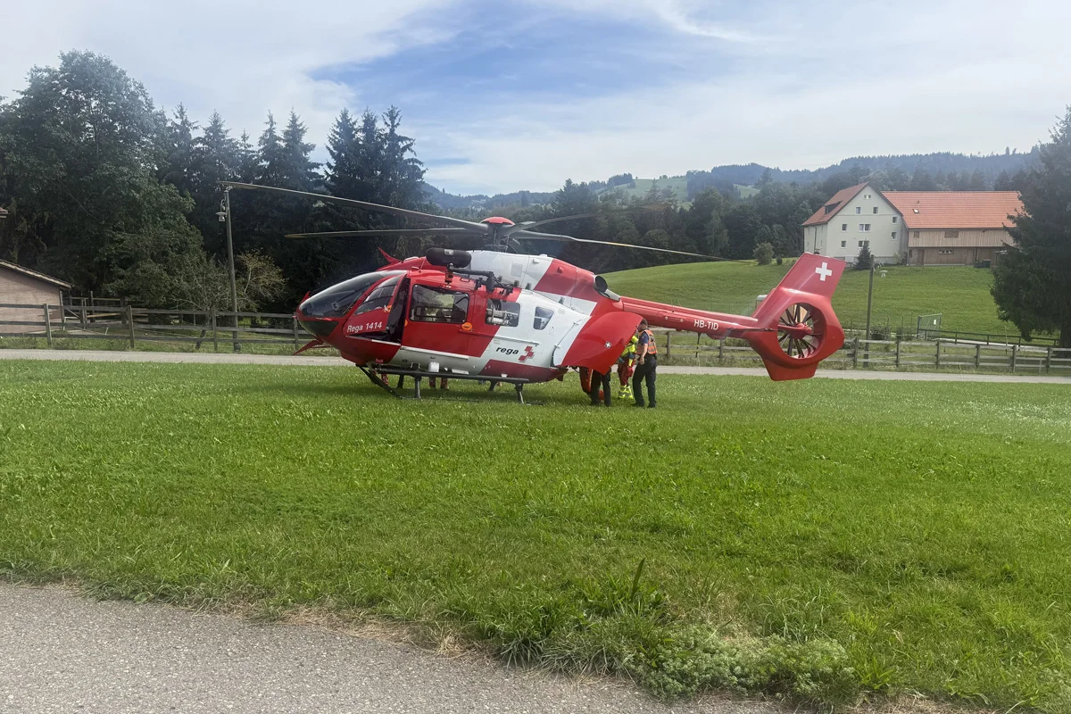 Ein Rega-Hubschrauber steht auf einer Wiese, umgeben von Einsatzkräften. Im Hintergrund sind Wälder und Häuser sichtbar.
