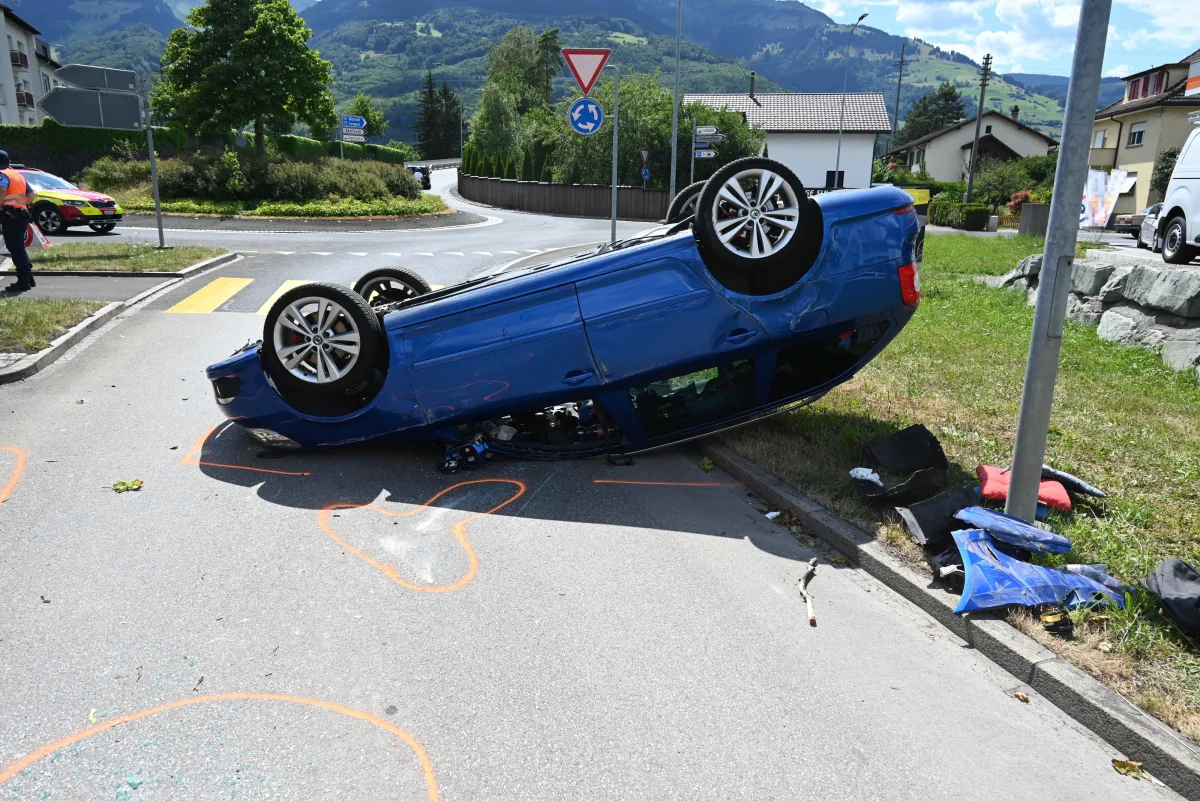 Auf der Straße liegt ein blauer Pkw auf dem Dach in der Nähe eines Kreisverkehrs. Teile des Fahrzeugs sind verstreut auf dem Gehweg.