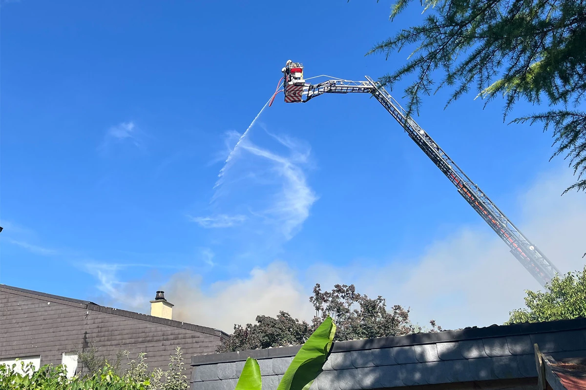 Ein Feuerwehrmann spritzt von einer ausgefahrenen Drehleiter aus Wasser auf ein Hausdach. Im Hintergrund ist blauer Himmel mit wenigen Wolken zu sehen, und Rauch steigt neben dem Gebäude auf.