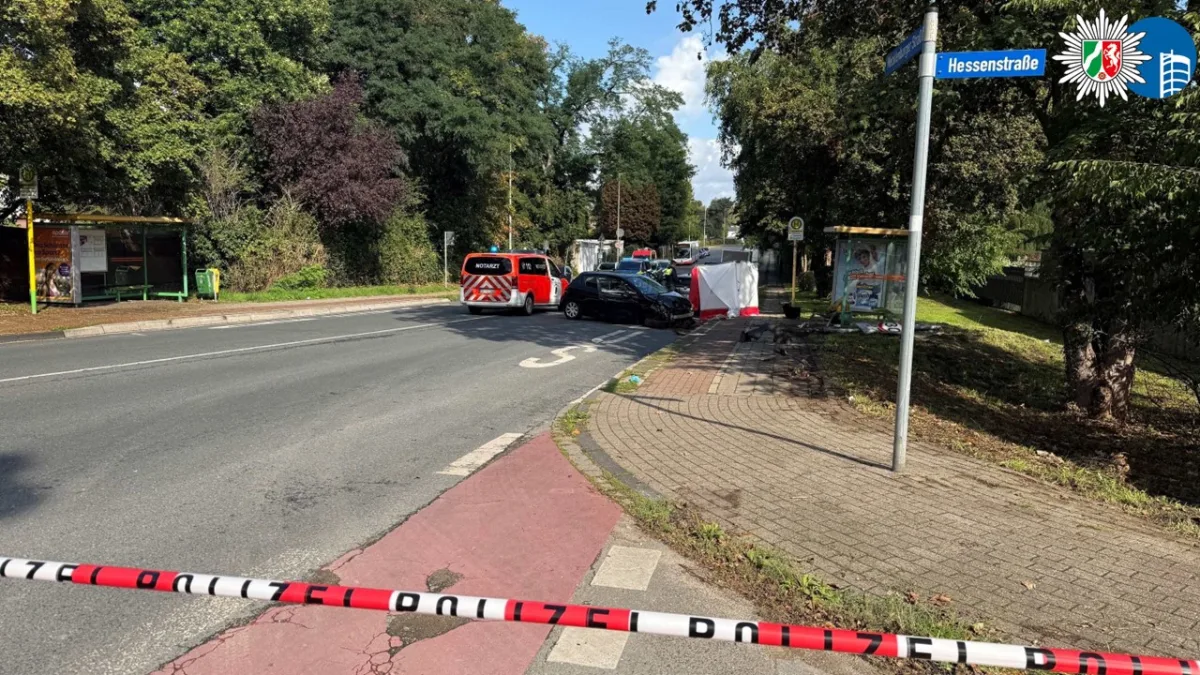 Die Szene zeigt einen Verkehrsunfall auf der Hessenstraße. Ein schwarzes Auto ist beschädigt, ein Rettungswagen ist vor Ort. Ein Polizeiband sperrt die Unfallstelle ab.