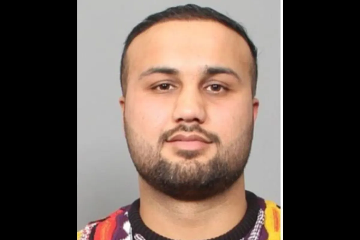Ein Porträtfoto zeigt eine Person mit dunklem Haar und Bart vor einem neutralen grauen Hintergrund. Die Person trägt ein bunt gemustertes Oberteil.