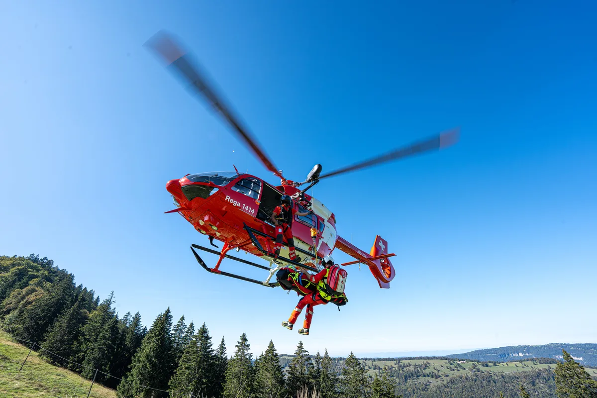 Ein roter Rega-Hubschrauber schwebt über einer Waldlichtung, während zwei Rettungskräfte am Seil arbeiten. Einer der Retter trägt eine Trage.