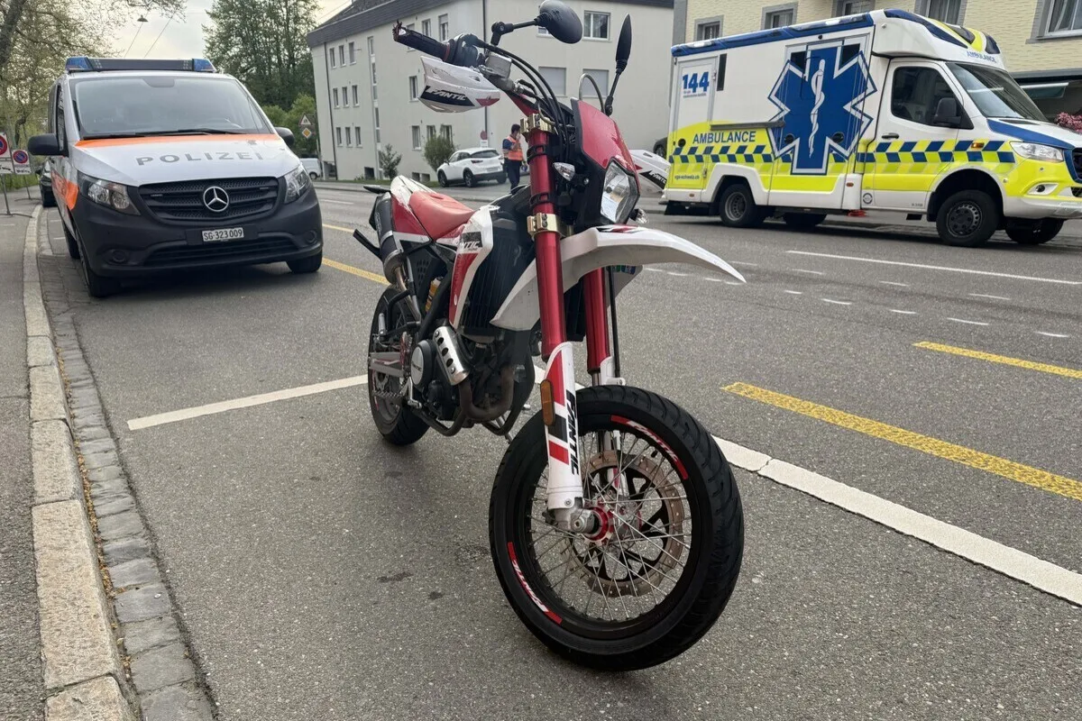 Ein Motorrad steht auf einer Straße, daneben sind ein Polizeiwagen und ein Krankenwagen zu sehen. Die Szene spielt in einer städtischen Umgebung.